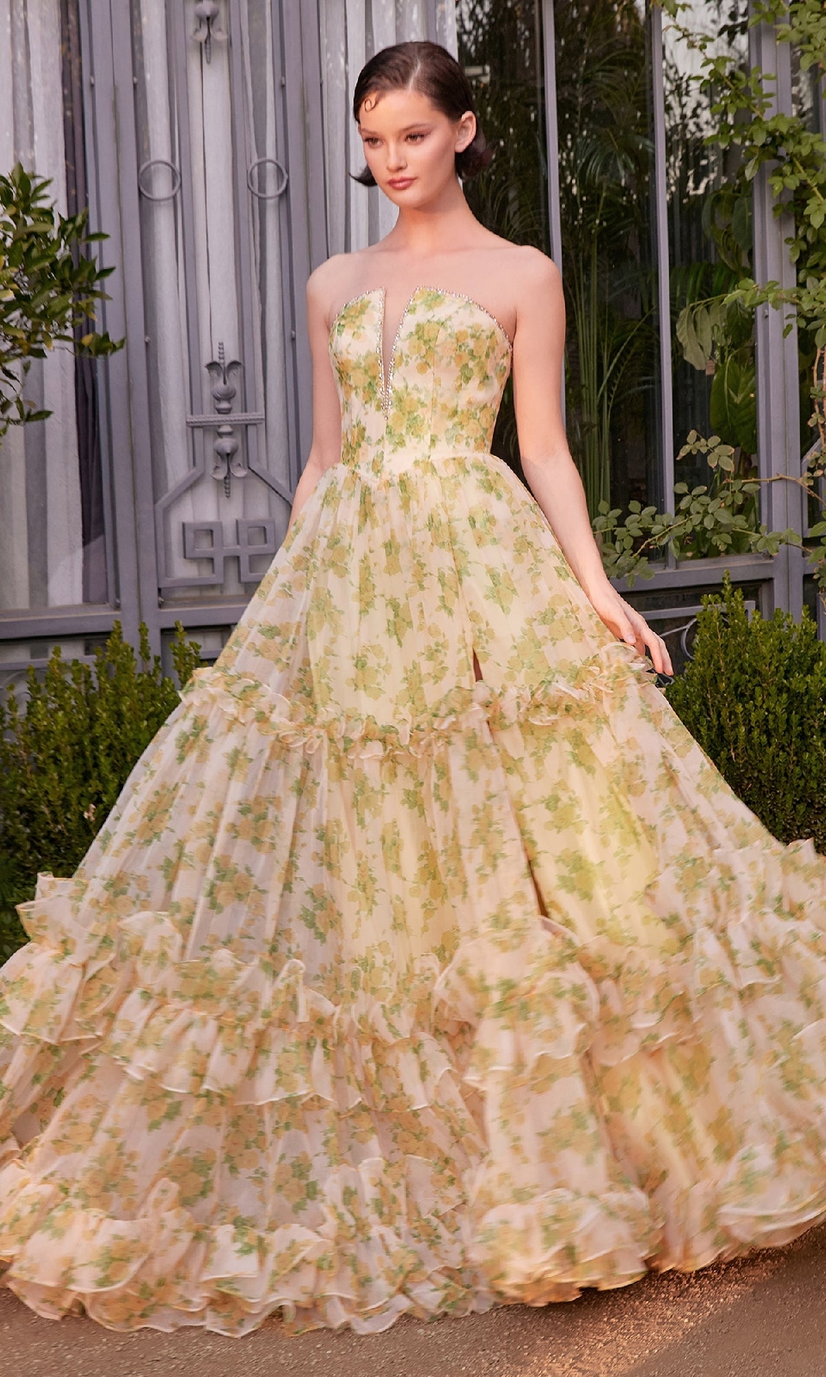 A woman stands outdoors before a building with lush greenery, wearing the Andrea & Leo A1430 Yellow Flora-Print Prom Gown, featuring ruffled layers and a strapless design.